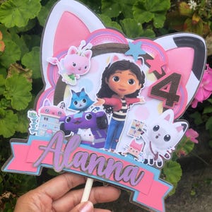 Könnte beinhalten: Bunter Tortenaufsatz mit einem Cartoon-Mädchen, Katzenfiguren, der Zahl 4 und dem Namen "Alanna" auf einem rosa Banner. Eine festliche Dekoration für eine Kindergeburtstagsparty mit Katzen-Thema.