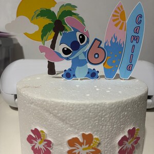 May include: A cake with a tropical theme, featuring a blue cartoon character holding a number 6. The cake is decorated with a yellow sun, palm tree, and two surfboards with the name "Camila". Pink and orange hibiscus flowers adorn the base.