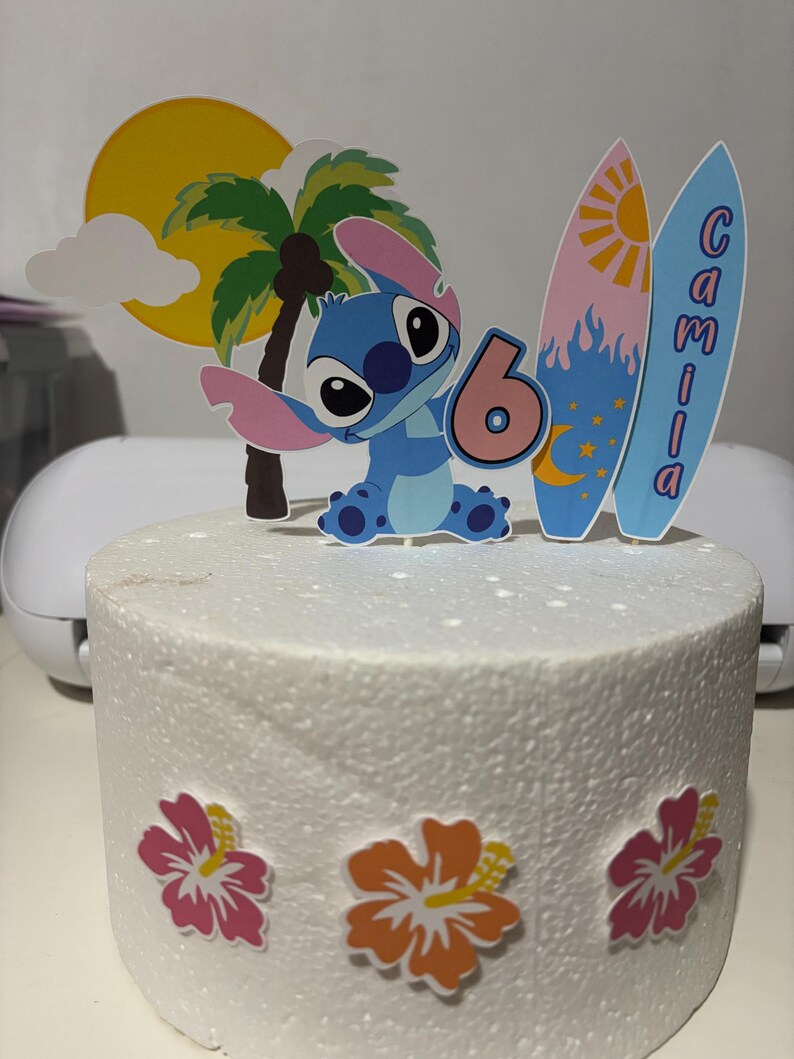 May include: A cake with a tropical theme, featuring a blue cartoon character holding a number 6. The cake is decorated with a yellow sun, palm tree, and two surfboards with the name "Camila". Pink and orange hibiscus flowers adorn the base.