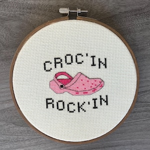 May include: A cross-stitch embroidery hoop featuring the words "CROC'IN ROCK'IN" above and below a pink clog with star details. The embroidery is set in a wooden hoop, perfect for wall decor.