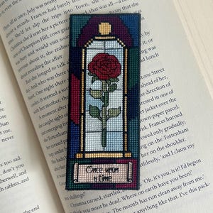 Puede incluir: Marcapáginas bordado con una rosa roja en un diseño de vidriera. El marcapáginas tiene forma rectangular con una pancarta que dice "Once Upon a Time". El fondo es una mezcla de azul, verde y rojo.
