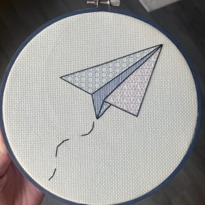 Könnte beinhalten: Gesticktes Papierflugzeug-Design in einem runden Rahmen. Das Papierflugzeug ist in Blau- und Grautönen gehalten, mit einer gepunkteten Spur. Der Hintergrund ist ein hellgrauer Stoff, und der Rahmen ist dunkelblau.