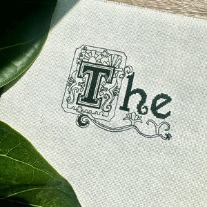 May include: Cross-stitch embroidery on white fabric. The design features the word "The" in black thread. The letter "T" is in a decorative frame with floral and geometric elements. The word "the" is in a simple font.