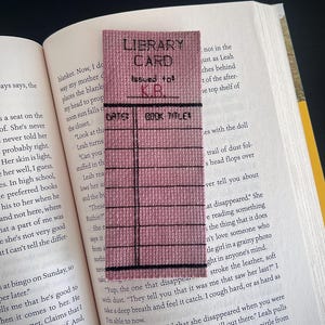 May include: A handmade bookmark with a library card design. The bookmark is a rectangular shape with a pink fabric background and black stitched lines and text. The text reads "LIBRARY CARD" and "Issued to: KB". The bookmark is placed inside an open book.