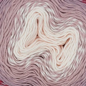 May include: A close-up of a ball of yarn with a gradient of colors. The yarn transitions from a deep burgundy at the outer edge, through shades of pink and mauve, to a creamy white center. The yarn is tightly wound.