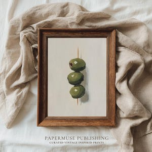 May include: A framed print depicting three green olives skewered on a cocktail stick. The artwork is set against a neutral background and framed in a dark wood. The print has the text "PAPERMUSE PUBLISHING" and "CURATED VINTAGE INSPIRED PRINTS".