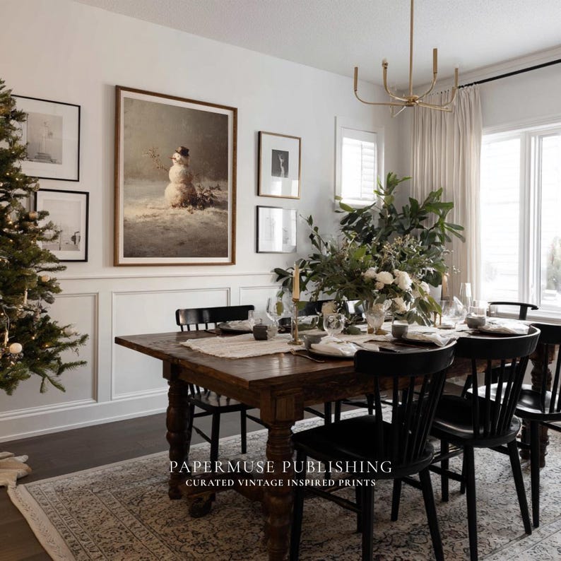 May include: A dining room scene featuring a dark wood table set for a meal, with black chairs and a floral centerpiece. A framed winter landscape painting of a snowman hangs on the wall, along with other framed art. The text "PAPERMUSE PUBLISHING CURATED VINTAGE INSPIRED PRINTS" is visible.
