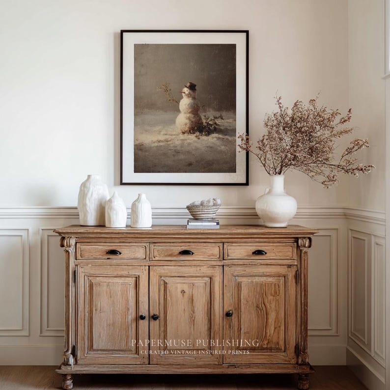 May include: A framed print of a snowman in a snowy landscape hangs above a wooden cabinet. The cabinet has three drawers and two doors. White vases and a decorative bowl sit on top of the cabinet. A white vase holds dried flowers.