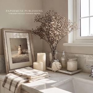 May include: A framed vintage print of a snowman in a snowy landscape, with the text "PAPERMUSE PUBLISHING CURATED VINTAGE INSPIRED PRINTS". The print is displayed on a marble countertop with candles, towels, and a vase of dried flowers.