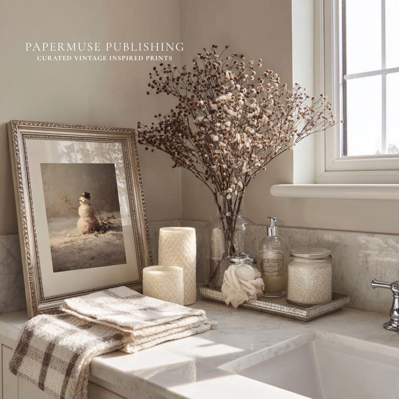 May include: A framed vintage print of a snowman in a snowy landscape, with the text "PAPERMUSE PUBLISHING CURATED VINTAGE INSPIRED PRINTS". The print is displayed on a marble countertop with candles, towels, and a vase of dried flowers.