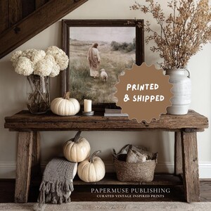 May include: A rustic wooden console table displays a framed print, white pumpkins, and floral arrangements. The print depicts a figure with a lamb. A woven basket and a gray throw blanket add texture. The text "PRINTED & SHIPPED" is visible.