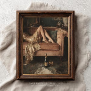 May include: Framed vintage print depicting a woman in a gold dress reclining on a pink sofa. A bottle of champagne and a glass are in the foreground. The print has a dark brown wooden frame.