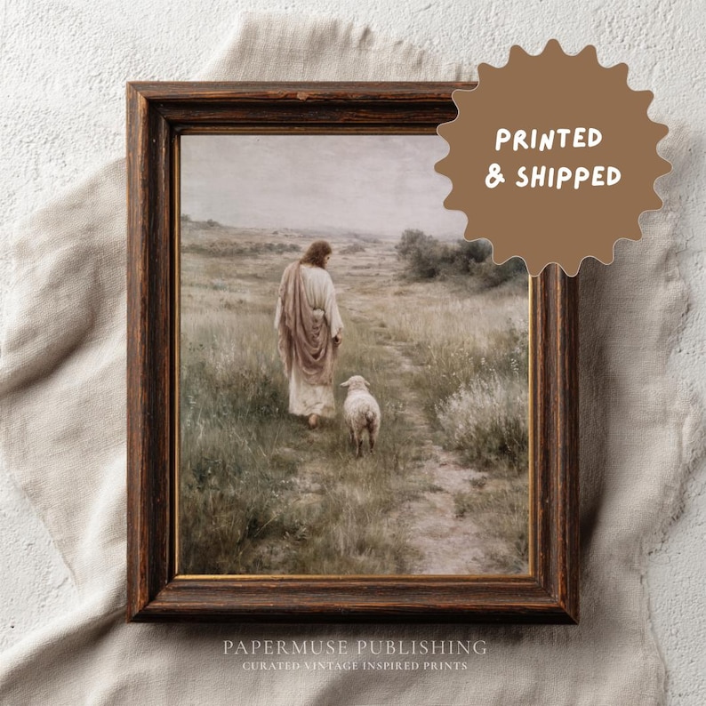 May include: A framed print of a religious scene. The artwork depicts a figure in flowing robes walking with a lamb on a path through a field. The frame is dark brown. The print has a vintage aesthetic.