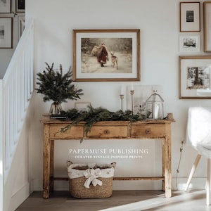 Puede incluir: Una consola de madera r&uacute;stica con tres cajones, decorada con una impresi&oacute;n enmarcada, vegetaci&oacute;n, velas y una linterna decorativa. Debajo hay una cesta tejida con un forro floral. La impresi&oacute;n enmarcada representa a una persona y un ciervo en un paisaje nevado. El texto "PAPERMUSE PUBLISHING" es visible.