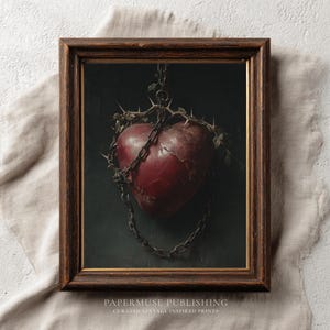 May include: A framed print featuring a dark red heart pierced with thorns and bound by a chain. The heart is set against a dark background, and the print is in a brown wooden frame. The print is from Papermuse Publishing.