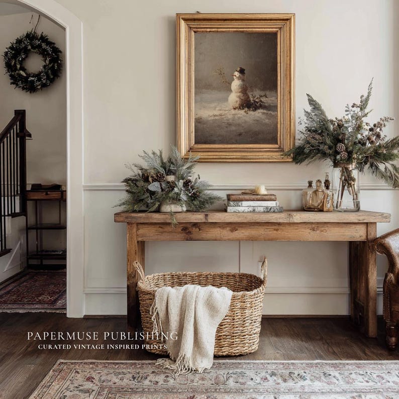 May include: A rustic wooden console table with a framed vintage snowman print, floral arrangements, and a woven basket with a cream-colored throw. A wreath hangs on the wall. The image is from Papermuse Publishing.