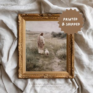 May include: A framed vintage print depicts a figure in flowing robes walking with a lamb in a grassy field. The artwork is in a gold ornate frame. The print has the text "PRINTED & SHIPPED" and "PAPERMUSE PUBLISHING CURATED VINTAGE INSPIRED PRINTS".