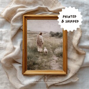 May include: A framed print with a gold-colored frame. The artwork depicts a person in a robe walking with a lamb on a path through a field. The text "PRINTED & SHIPPED" is in a white circle. The text "PAPERMUSE PUBLISHING" is at the bottom.