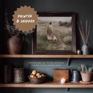 May include: A framed print of a religious scene depicting a figure with a lamb, displayed on a wooden shelf with decorative items. The print is in a dark wooden frame. The text "PRINTED & SHIPPED" is in a brown circle. The text "PAPERMUSE PUBLISHING" is below the shelf.