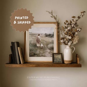 May include: A framed print of a religious figure with a lamb, displayed on a wooden shelf with books and a vase of dried flowers. The print is in a light-colored frame. The text "PRINTED & SHIPPED" is in a brown circle. The text "PAPERMUSE PUBLISHING" is below the shelf.
