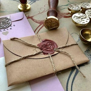 May include: A brown paper envelope sealed with a red wax stamp and twine. A wooden handled gold wax stamp with a heart design is nearby, along with other wax stamps and envelopes. The envelopes are tied with twine.