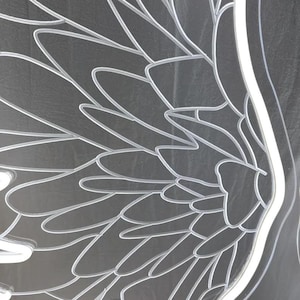 May include: A clear acrylic panel featuring a detailed laser-etched design of large, layered bird wings. The etched lines are white and create a high-contrast pattern against the transparent surface, showing individual feathers in a fan-like arrangement.