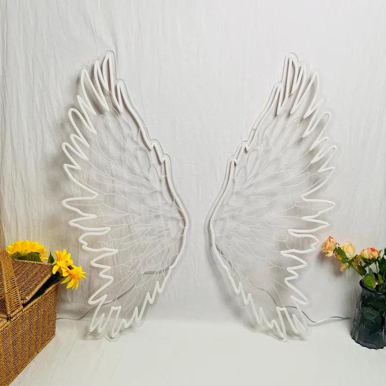 May include: A pair of large white neon angel wings mounted on a wall. To the left sits a woven wicker basket containing yellow flowers, and to the right is a glass vase holding a bouquet of peach and yellow flowers with green leaves.