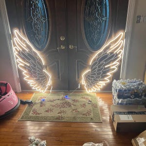 May include: A pair of illuminated LED neon-style wall signs shaped like large feathered angel wings. The signs emit a warm white glow and are mounted on a dark wooden double door with oval glass panels, featuring detailed feather outlines.