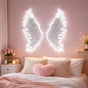 May include: A pair of illuminated white neon angel wings mounted on a pink wall above a bed. The wings feature detailed feather patterns outlined in bright white light, creating a glowing effect against the solid pink background.
