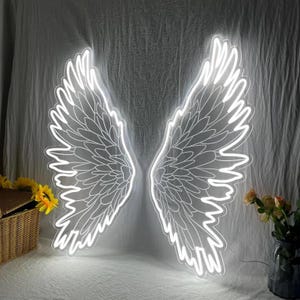 May include: A large, bright white LED neon sign shaped like a pair of expansive, detailed angel wings. The glowing light fixture features intricate feather patterns and is mounted against a plain, draped fabric backdrop.