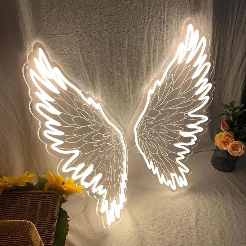 May include: A bright white LED neon sign shaped like a pair of large, detailed angel wings. The wings feature intricate feather outlines and emit a soft, glowing light, creating a distinct silhouette against the textured fabric backdrop.