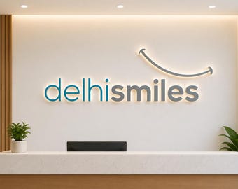 May include: A wall-mounted 3D sign displaying the text delhismiles in blue and gray lettering with a backlit smile graphic above the letters. The sign is positioned on a white wall above a white reception desk with two green potted plants.