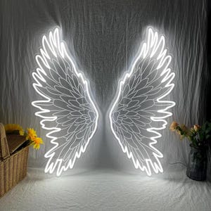 May include: A bright white LED neon sign shaped like a pair of large, detailed angel wings. The wings feature intricate feather outlines and are mounted against a textured white fabric backdrop, flanked by a woven basket and a glass vase.
