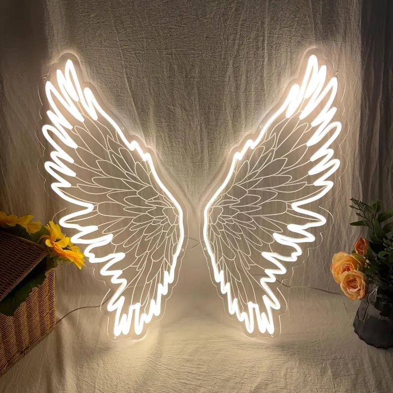 May include: A pair of bright white LED neon angel wings mounted on a clear backing. The light fixture features detailed feather outlines and a symmetrical design, creating a glowing silhouette against the fabric backdrop.