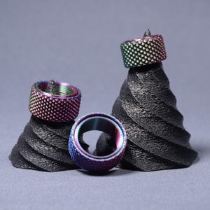 Könnte beinhalten: Drei schillernde Ringe mit strukturierter Oberfläche werden auf schwarzen, spiralförmigen Ständern präsentiert. Die Ringe wechseln die Farbe und zeigen Farbtöne von Lila, Blau und Grün. Die Ständer bilden einen kontrastierenden Hintergrund und heben die leuchtenden Farben der Ringe hervor.