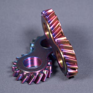 Könnte beinhalten: Zwei schillernde Metallzahnräder mit Regenbogen-Finish, die Farbtöne von Rosa, Lila und Gold zeigen. Die Zahnräder sind ineinandergreifend, mit sichtbaren Zähnen und kreisförmigen Öffnungen, vor grauem Hintergrund.