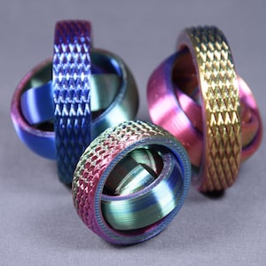 Könnte beinhalten: Drei schillernde, geometrische Fidget-Spielzeuge mit strukturierten Bändern und Innenringen. Die Farben wechseln von Blau über Lila zu Gold und erzeugen einen dynamischen visuellen Effekt. Die Spielzeuge sind mit komplizierten Mustern gestaltet.