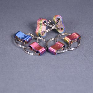 Könnte beinhalten: Nahaufnahme mehrerer Schlüsselanhänger mit schillernden Bändern. Die Schlüsselanhänger haben silberfarbene Metallringe und die Bänder zeigen eine Reihe von Farben, darunter Blau, Rosa und Gelb. Die Schlüsselanhänger sind auf einer grauen Oberfläche angeordnet.