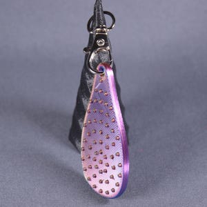Könnte beinhalten: Ein tropfenförmiger Schlüsselanhänger mit einem lila-rosa Banddesign, verziert mit kleinen, kupferfarbenen Perlen. Der Anhänger ist an einem silberfarbenen Verschluss und einer schwarzen, strukturierten Basis befestigt.
