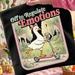 Puede incluir: Bolso tote negro con un dibujo de un ganso en un scooter, sosteniendo un café. La imagen tiene un borde rosa y verde y el texto "Off to Regulate Emotions". El ganso lleva un lazo rosa. El diseño es de estilo retro.