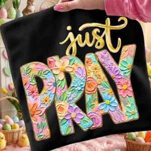 May include: Black pillowcase with the words "just PRAY" in gold script and floral letters. The letters are filled with colorful flowers in shades of pink, orange, blue, and purple. The pillowcase is held up by a person.