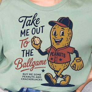 Puede incluir: Camiseta verde salvia con un personaje de cacahuete de dibujos animados con uniforme de béisbol, sosteniendo una pelota de béisbol y usando un guante. La camiseta tiene el texto "Take Me Out To The Ballgame" y "Buy Me Some Peanuts and Crackerjacks".