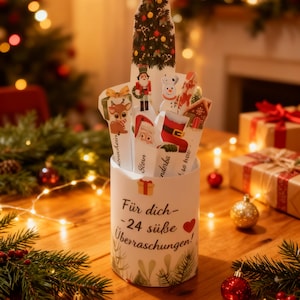 Pode incluir: Um calendário do advento cilíndrico branco com uma árvore de Natal e recortes de personagens festivos. O calendário tem o texto "Für dich - 24 süße Überraschungen!" e é decorado com enfeites de Natal e vegetação.