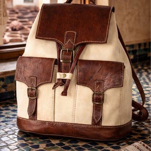 Puede incluir: Mochila de cuero crema y marrón con dos bolsillos delanteros y cierre de solapa. La mochila presenta detalles, hebillas y correas de cuero marrón. La bolsa tiene un asa superior y correas ajustables.