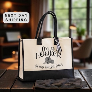 May include: Cream-colored tote bag with black trim and handles. The bag features the text "I'm a Hooker in my spare time" with a yarn ball graphic. A black and white striped ribbon is tied to the handle.
