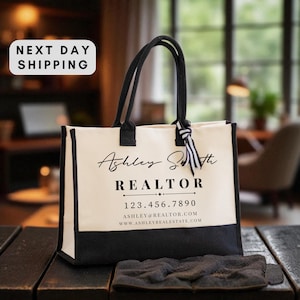 May include: Cream-colored tote bag with black accents, featuring the name "Ashley Smith" and the title "REALTOR" in elegant script. Includes a phone number and website address. The bag has black handles and a black bottom.
