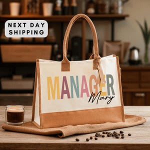 May include: Cream-colored tote bag with brown accents and handles. The bag features the word "MANAGER" in colorful letters and the name "Mary" in a script font. The bag is sitting on a brown surface.