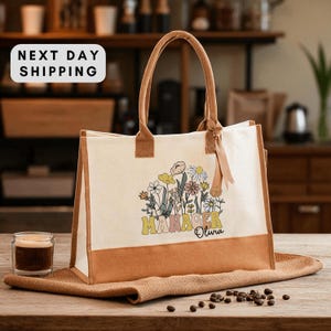 May include: Cream-colored tote bag with brown accents and handles. The bag features a floral design with the word "MANAGER" and the name "Olivia" in a retro font. The bag is sitting on a brown surface.