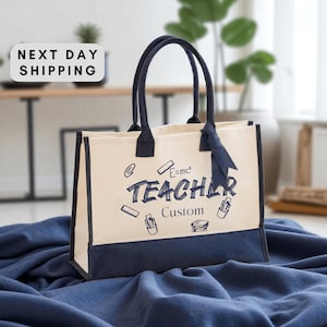May include: Cream-colored tote bag with navy blue accents and handles. The bag features the word "TEACHER" in navy blue script, along with custom text and small illustrations. A navy blue ribbon is tied to one handle.