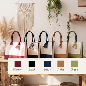 A row of five colorful tote bags with handles, each in a different color, displayed on a wooden table. The bags are arranged in a row, with the first bag being maroon, the second black, the third navy, the fourth coffee, and the fifth green. The background features a wall with a macrame wall hanging and potted plants.